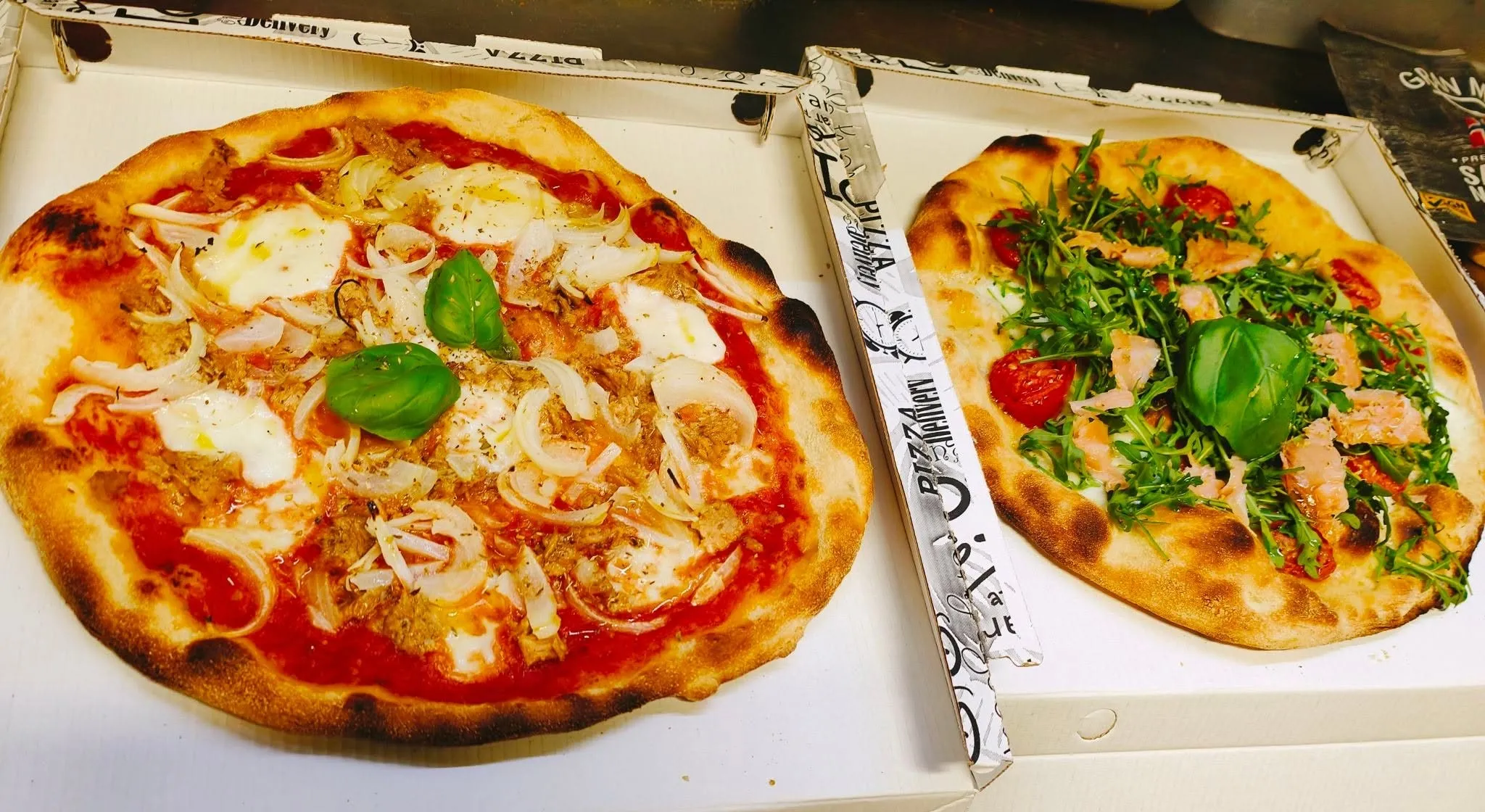Due pizze pronte nei cartoni da asporto, una tonno e cipolla e una salmone e rucola